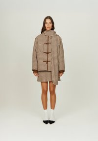 Καφέ και κρεμ καρό εξωτερικό ένδυμα με ψηλό γιακά, που διαθέτει κλείσιμο με κουμπί και ασύμμετρο σχέδιο, συνδυασμένο με φούστα στον ίδιο ύφασμα.