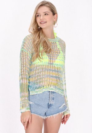 Ung kvinde med langt blondt hår iført en neon gul bikinitop, flerfarvet gennemsigtig strikket sweater og lyseblå denimshorts, som smiler let.