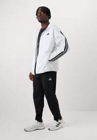 Casaco leve branco com riscas pretas nas mangas, calças de desporto pretas e tênis cinzentos com detalhes brancos. Logotipo da Adidas visível.