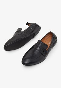 Los zapatos negros de piel tipo mocasín presentan un acabado suave, diseño de punta afilada y una tira decorativa en la parte superior. Forro texturizado visible en el interior.