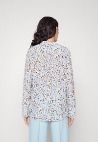 Blumenbluse aus hellblauem Stoff mit fließendem Design, das abstrakte Muster in Orange und Lila zeigt, und einem knopfartigen Detail auf der Rückseite.