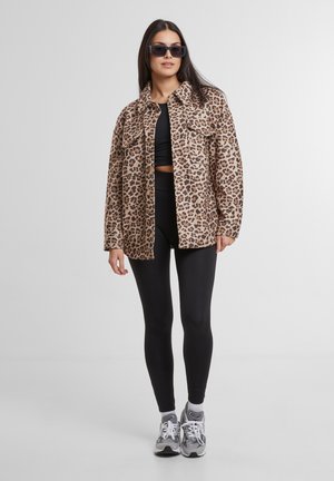 Moteris nešiojanti juodus saulės akinius, striukę su leopardiniu raštu, juodą trumpą palaidinę, juodas tamprės ir pilkus sportinius batelius, stovinti prieš paprastą foną.