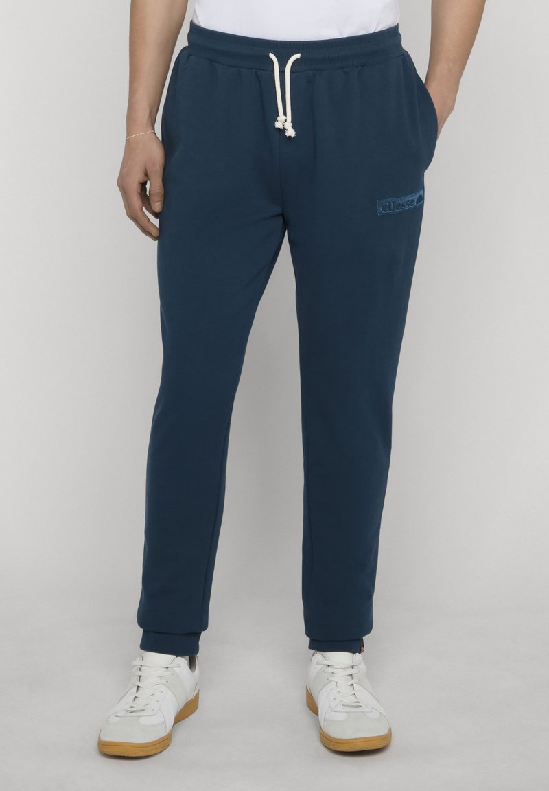 Personne portant un pantalon de survêtement bleu marine avec un cordon blanc et des baskets blanches à semelles en gomme, debout devant un fond uni.