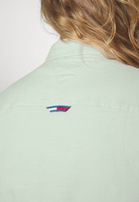 Ljusgrön kragskjorta med broderad röd, vit och blå logotypdetalj på baksidan, med en slät textur och prydlig sömnad.