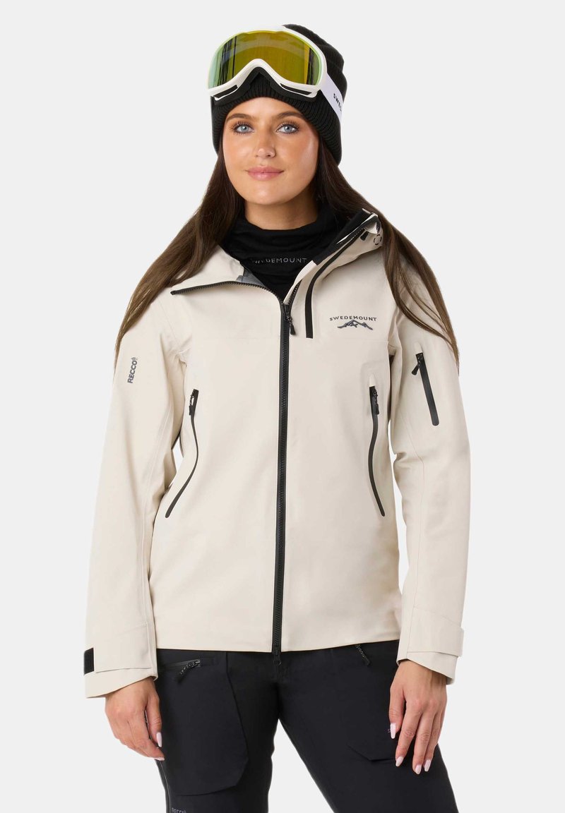 Giacca impermeabile beige con zip e dettagli neri, dotata di chiusura frontale a zip, tasche laterali e cappuccio. Indossata con occhiali da sci e un berretto.