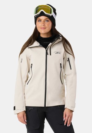 Beige waterproof jacket with black zippers and accents, featuring a zip-up front, side pockets, and a hood. Worn with ski goggles and a beanie.