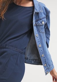 Personne portant une robe bleu marine avec des poches et une veste en denim partiellement enlevée avec des boutons en argent, sur un fond uni.