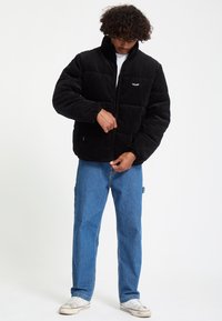 Homme aux cheveux bouclés portant une doudoune noire qu’il ferme, un jean bleu et des baskets blanches sur un fond blanc uni.