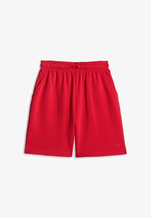 Garçon debout, les mains sur les hanches, regardant sur le côté, portant un t-shirt noir à manches courtes, un short rouge, des chaussettes blanches et des baskets noires et blanches.