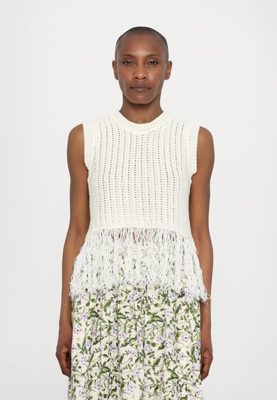 Strikket ærmeløs top i creme med et tekstureret design og en frynsekant. Båret over en blomsterprint nederdel med lilla blomster og grønne blade.