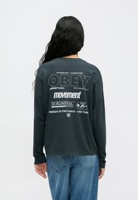 Czarny t-shirt z długim rękawem z białym graficznym napisem na plecach, o luźnym kroju i miękkiej fakturze. W zestawie z jasnoniebieskimi dżinsami.