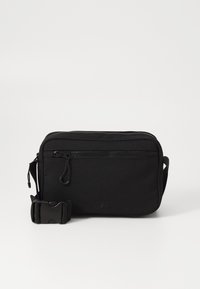 Black fabric crossbody bag with a zippered main compartment, front pocket, and adjustable strap. Textured surface and logo detail visible.