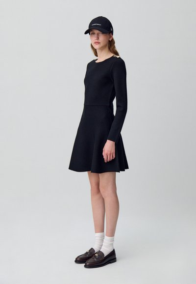 Abito nero a maniche lunghe con gonna svasata e dettagli dorati sulle spalle, abbinato a un berretto nero e mocassini scuri indossati sopra calzini bianchi.