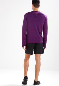 Fioletowa koszulka sportowa z długim rękawem o fakturowanym wzorze, w połączeniu z czarnymi szortami i czarnymi butami sportowymi, widziana z tyłu.