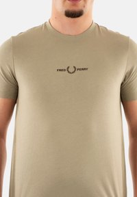 Camiseta de manga corta color caqui fabricada en algodón suave, con un logo bordado en marrón que presenta un diseño de laurel en el pecho.