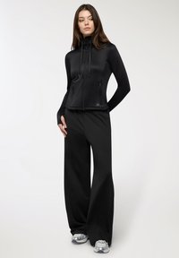 Giacca nera con zip e colletto alto, abbinata a pantaloni neri larghi. Il materiale sembra elegante e liscio, adatto per abbigliamento sportivo.