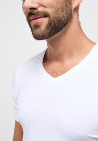 Hvid kortærmet t-shirt med V-udskæring, lavet af glat stof, tætsiddende silhuet, som viser en subtil tekstur ved udskæringen.
