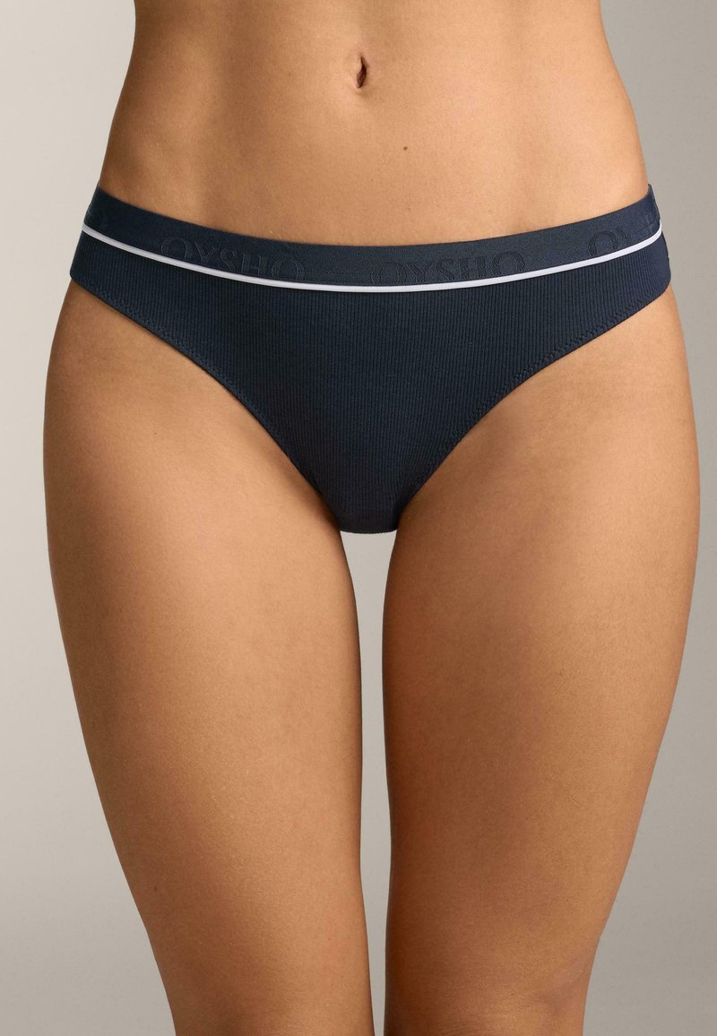 Slip bikini côtelé bleu marine avec une bande accent blanche et une ceinture élastique à logo, présentant une texture lisse et une coupe ajustée.