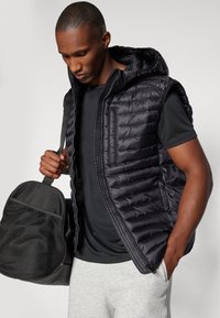 Črna podložena brezrokavnik s kapuco, z zadrgo in žepom na prsih. Model drži črno športno torbo, oblečen je v sivo spodnje oblačilo in črno zgornje oblačilo.