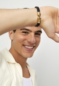 Pulsera de cuero negro con un cierre en tono dorado, llevada en una muñeca. El cierre tiene un acabado brillante, que complementa la textura suave del cuero.