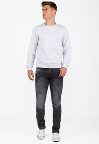 Felpa grigia chiara con scollatura a girocollo e polsini a coste, abbinata a jeans grigi scuri. Il modello è in piedi di fronte a uno sfondo bianco semplice.