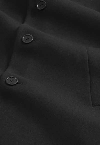 Mustapakka villakangas takki, jossa on sileä pinta, kaksi suurta mustaa nappia ja hienovarainen taskutaito edessä toiminnallista muotoilua varten.