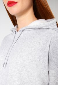 Mujer con largo cabello rojo viste una sudadera con capucha gris clara y cordones, labios pintados de un rojo brillante, contra un fondo sencillo.