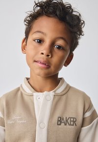 Veste de sport beige avec des accents blancs contrastants, boutons pression et logo "BAKER" imprimé en gras. Matière en coton doux, col rond.