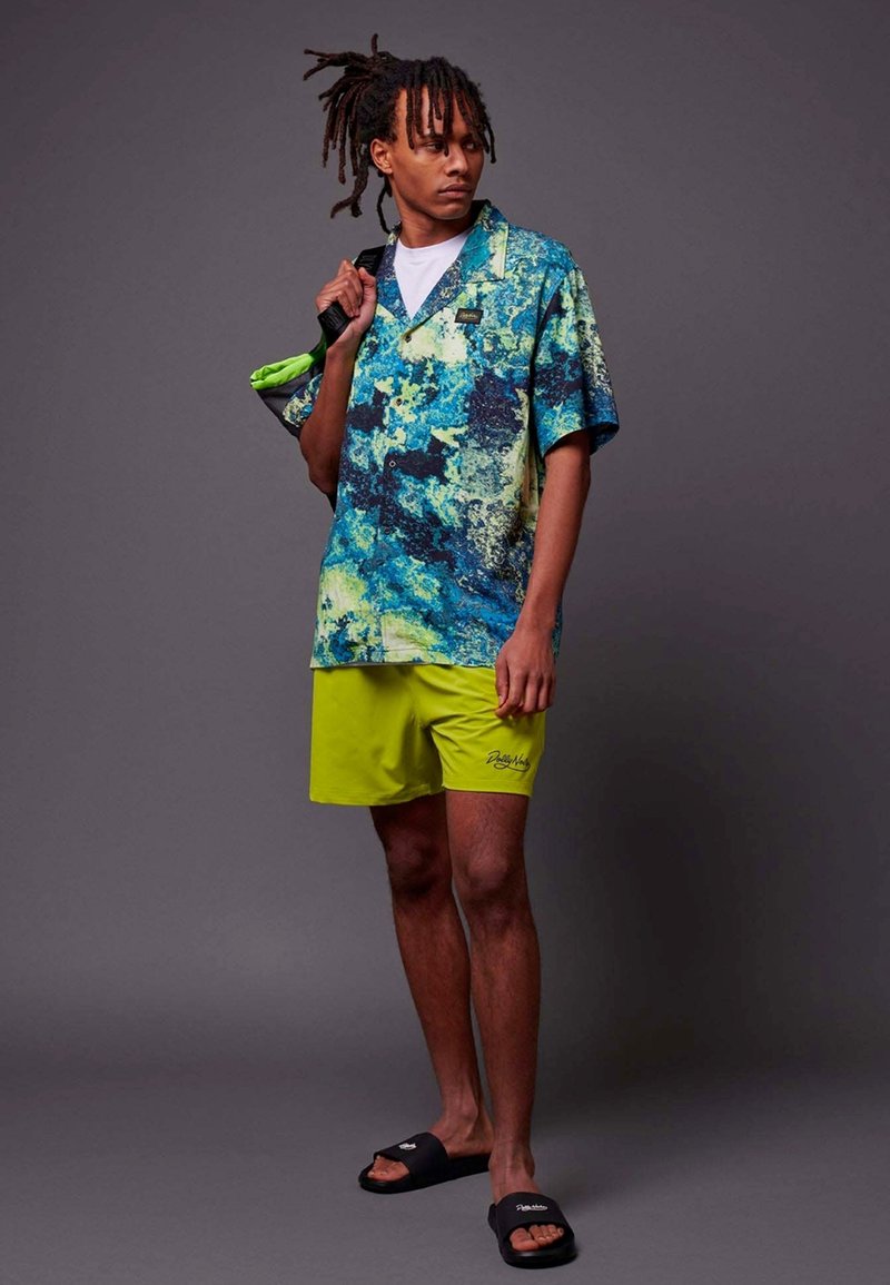 Chemise courte à manches colorées avec un motif abstrait bleu et vert, associée à un short jaune vif. Le modèle porte des sandales noires à semelle plate.