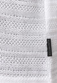 Hvid strikket stof med et åbent, tekstureret mønster. Har et lille sort label med "ALLSAINTS" i hvid skrift på kanten.