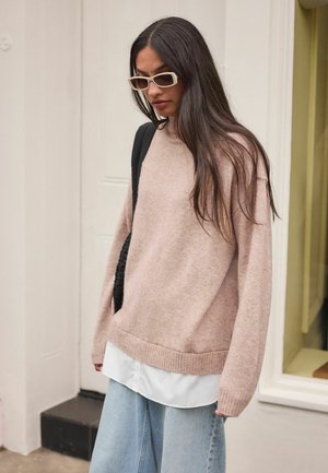 Maglione oversize rosa chiaro con orlo a costine, indossato sopra una camicia bianca con bottoni. Abbina con jeans a gamba larga blu chiaro e uno zaino nero.
