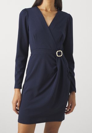 Femme portant une robe portefeuille bleu marine à manches longues avec un col en V et un détail de boucle de ceinture à la taille.