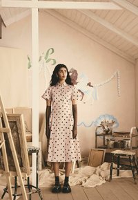 Vestido rosa con lunares, de mangas cortas, cuello y bolsillos. La modelo lleva guantes marrones y zapatos de plataforma negros, posicionada en un entorno de estudio.