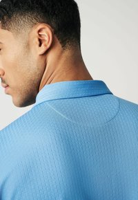 Hellblau gemustertes Polohemd mit Kragen, das ein dezentes Rautenmuster und einen glatten Stoff aufweist. Von hinten präsentiert, um die Details hervorzuheben.