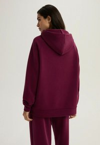 Burgundowa bluza z kapturem, o luźnym kroju, z obniżonymi ramionami i ściągaczami. Materiał wygląda na miękki i gładki w dotyku.