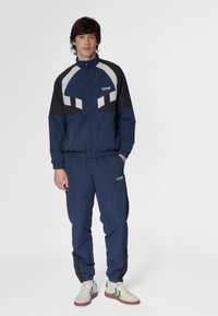 Marinblå träningsdräkt med en zip-up jacka med vita och svarta detaljer, matchande byxor och beige sneakers med gröna ränder.
