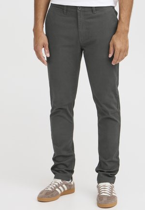 Chinos grises de corte slim, fabricados en algodón, con frente plano, pasadores para cinturón y bolsillos laterales, combinados con zapatillas deportivas grises y blancas.