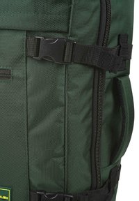 Mochila de tela verde con una superficie texturada, herrajes negros, cremallera lateral y correas ajustables con detalle de cierre tipo clip.
