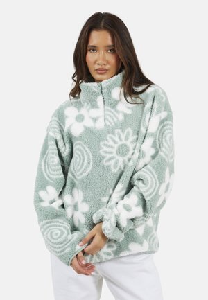 Donna con capelli lunghi castani che indossa un pile con motivo floreale verde e bianco e una cerniera a un quarto, in piedi davanti a uno sfondo bianco.