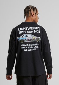 Juodas ilgarankovis marškinėliai su automobilių grafika ir balta tekstu nugaroje. Tekste yra "LIGHTWEIGHT 1991 UTM NO2" ir po juo esanti šūkis.