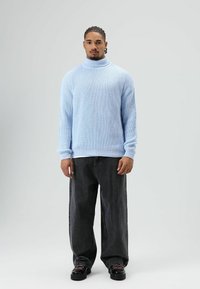 Pull à col roulé côtelé bleu clair associé à un jean noir ample et à des chaussures noires. Le pull présente un tricot texturé et des poignets ajustés.