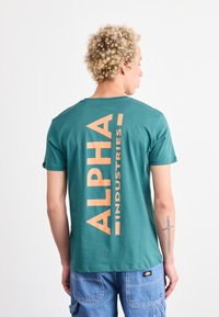 Turkizna majica s kratkimi rokavi z velikim oranžnim tiskom "ALPHA INDUSTRIES" na hrbtu. Izdelana iz mehkega bombažnega materiala, ima okroglo ovratnico.