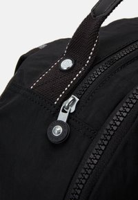 Musta reppu, jossa on nylonkangas, sujuva vetoketju, teksturoitu kahva, jossa on ommeltu yksityiskohta, ja pyöreä logo-aksentti.