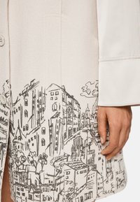 Cremfarbener Wollmischmantel mit einem handgezeichneten Stadtbild-Print in Schwarz am Saum, mit Knopfverschluss und breiten Bündchen.