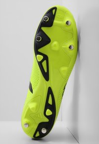 Jasně žlutá podrážka fotbalových kopaček se šesti kovovými hroty a černými gripovými vzory, zobrazená zdola na bílém pozadí.