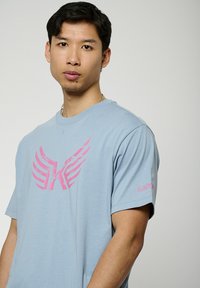 Lichtblauw T-shirt met korte mouwen, gemaakt van zachte stof, bevat een grote roze vleugelafbeelding op de voorkant en een roze logo op de mouw.