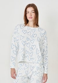 Mujer joven con cabello largo que lleva una blusa y pantalones blancos a juego, cubiertos de un patrón de corazones dibujados en azul, de pie contra un fondo blanco liso.