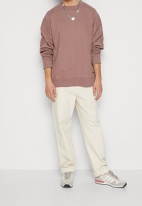 Braunes Oversize-Sweatshirt mit überschnittenen Schultern, cremefarbene, locker sitzende Hose und graue Sneaker mit dezenten roten Akzenten.