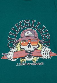 Tyrkysové tričko z látky s grafickou potlačou lebky s blond vlasmi, ktorá nosí slnečné okuliare a drží červenú surfovaciu dosku, spolu s textom "Quiksilver A Touch of Paradise."