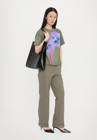 Olivenfarbener T-Shirt mit floralem Grafikdesign, kombiniert mit olivfarbenen Hosen und einer schwarzen Umhängetasche. Das Model trägt spitze schwarze Pumps.